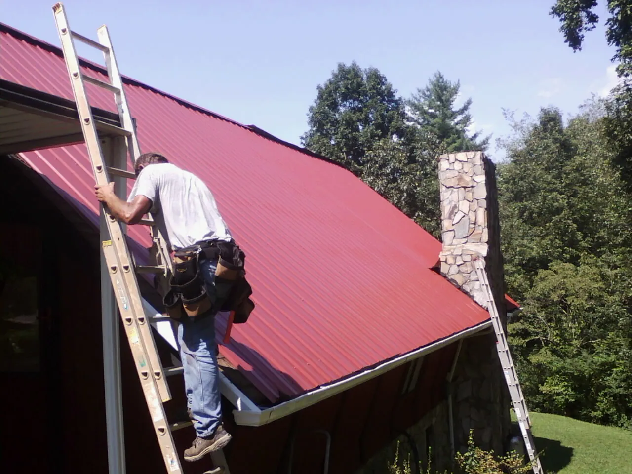 Roofer on a ladder beside a red metal roof and stone chimney — Soffit & Fascia Repair by Roofing Brothers in Emerson
